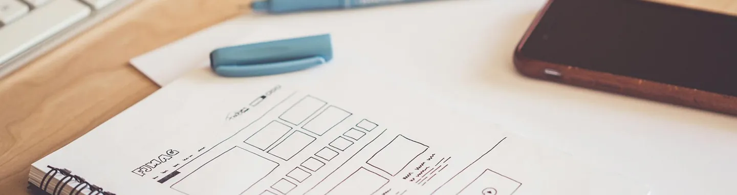 UI/UX Design Notebook with UI wireframe sketches next to a smartphone and keyboard